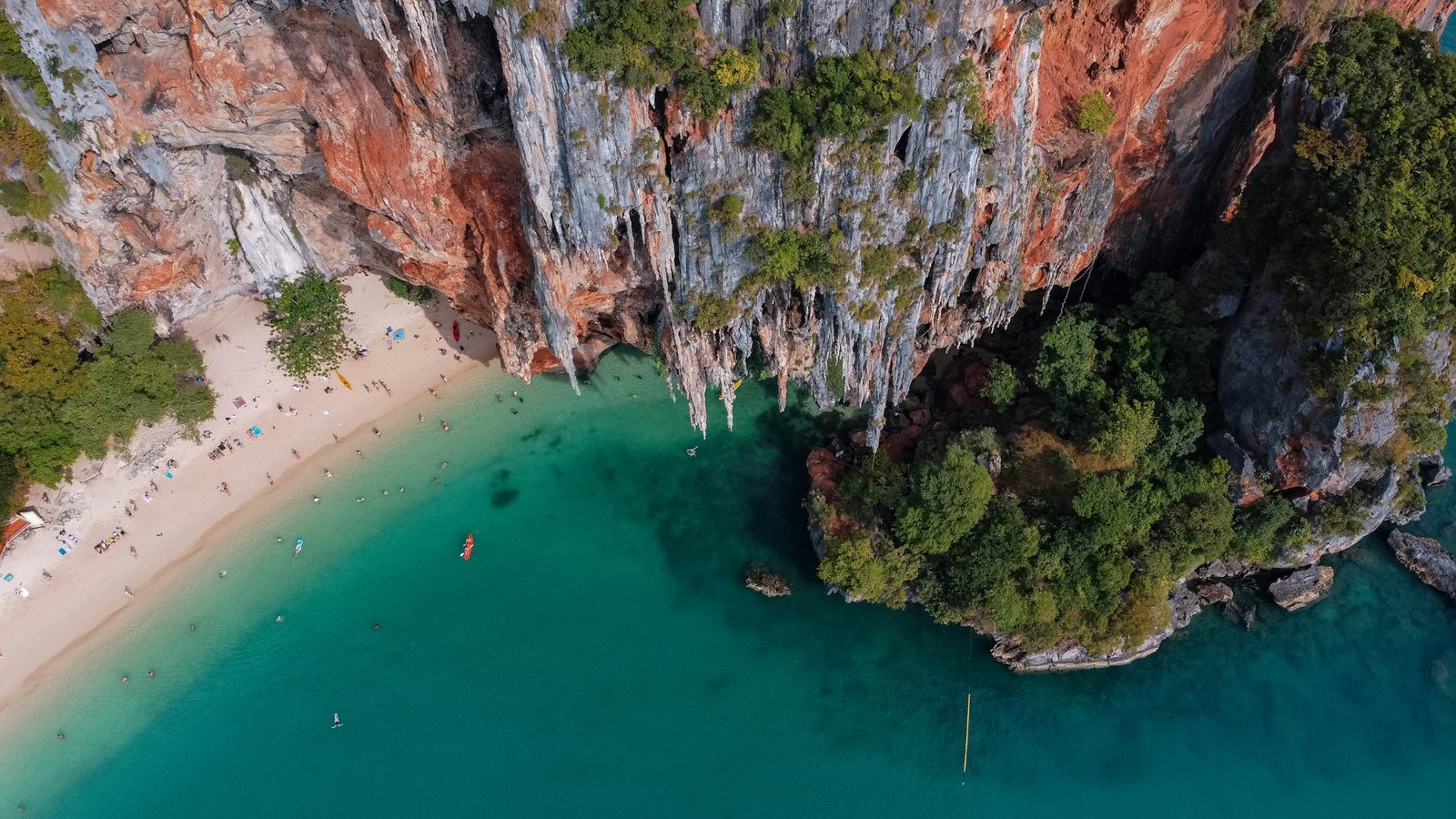 An aerial view of turquoise water meeting a white sand beach beneath towering limestone cliffs