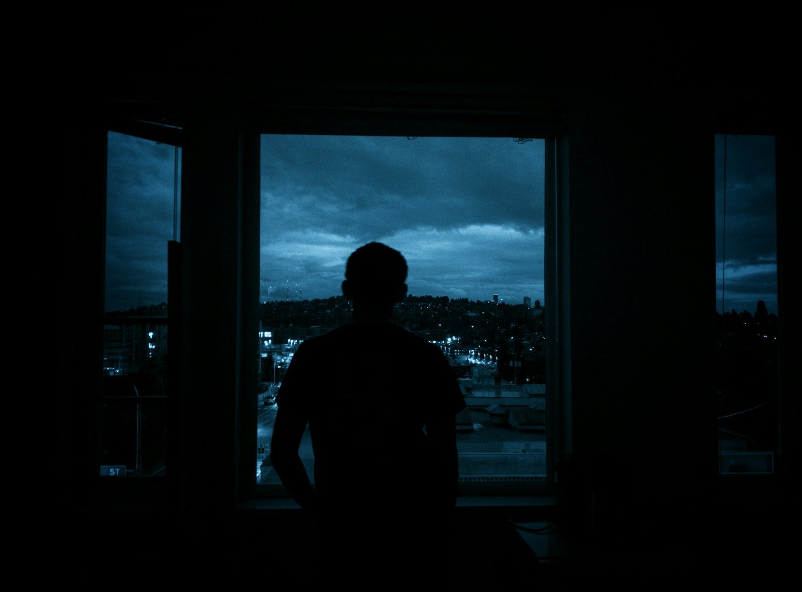 A solitary figure silhouetted against a dark window overlooking a quiet city at night